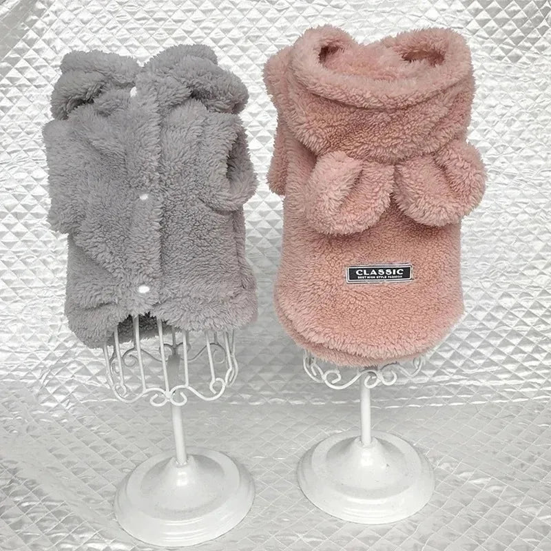 Two fluffy dog coats, one gray and one pink, on mannequin stands against a textured white background.