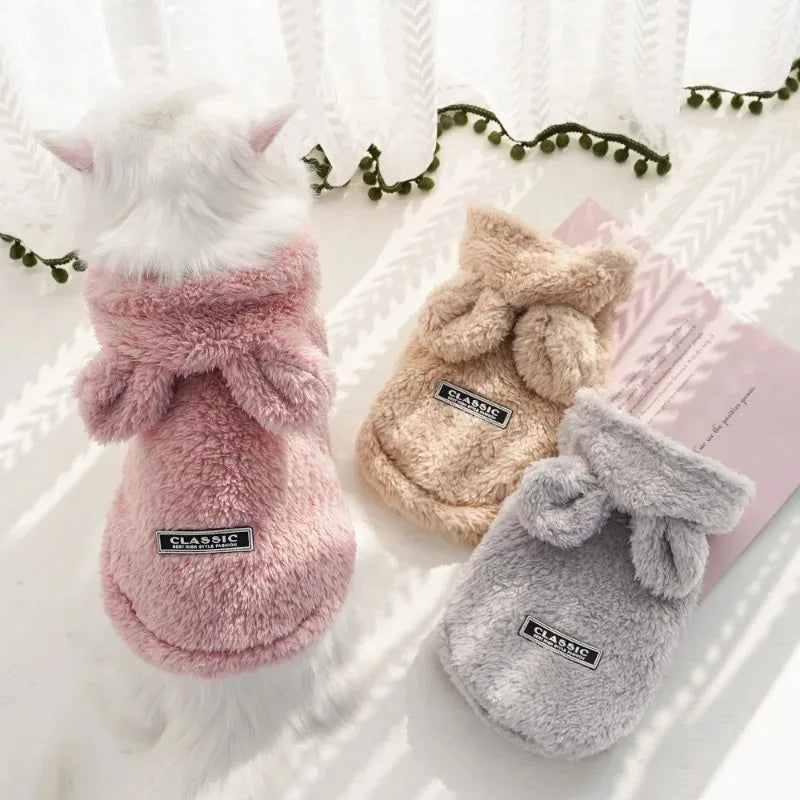 Three pairs of fluffy pet slippers in pink, beige, and gray on a white surface with a decorative background.